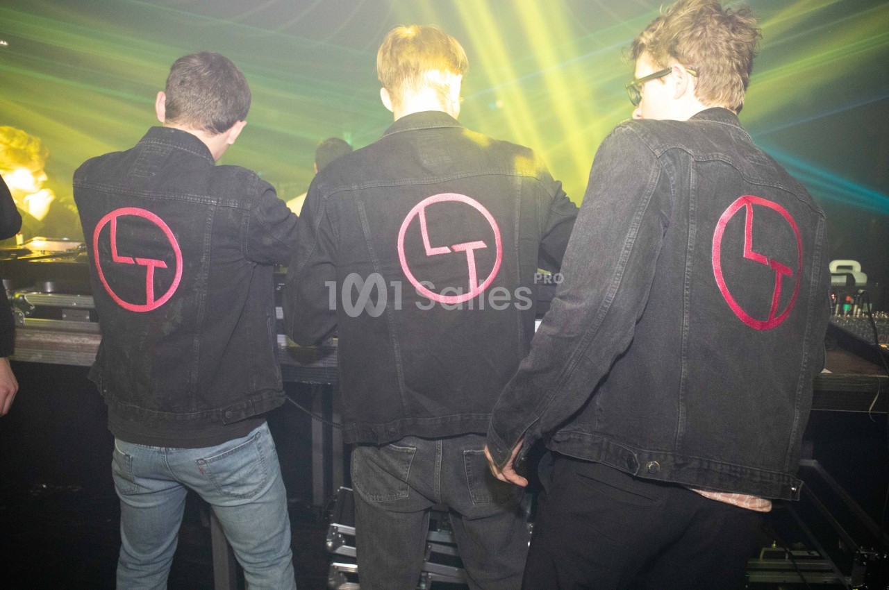 Trois personnes de dos portant des vestes en jean noir avec un logo rouge, dans une ambiance de lumière tamisée.