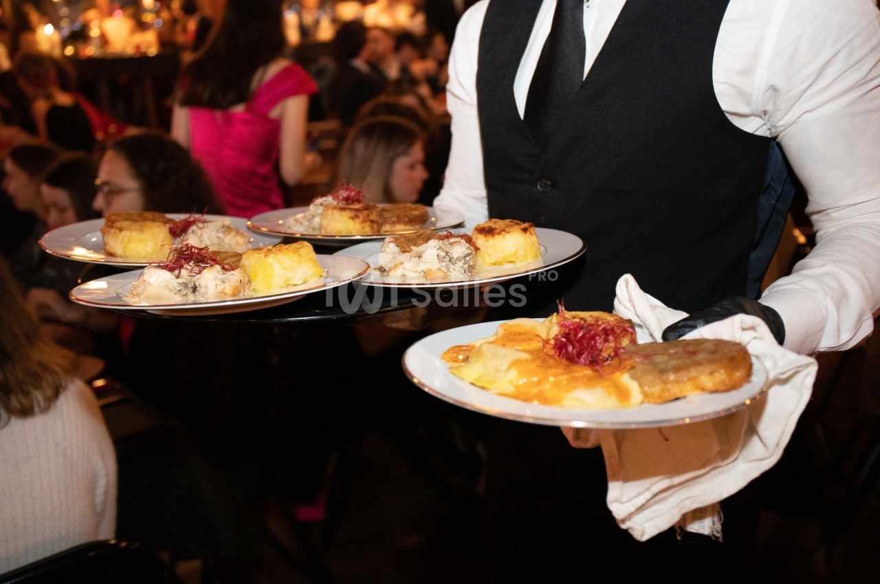 Un serveur en gilet noir porte plusieurs assiettes contenant des plats variés lors d'un événement en intérieur.