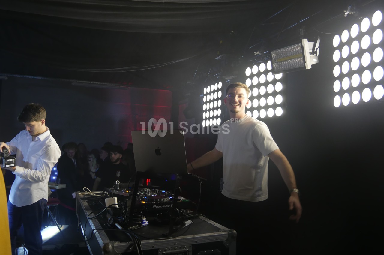 Un DJ souriant devant son équipement, éclairé par des projecteurs, dans une ambiance de soirée.