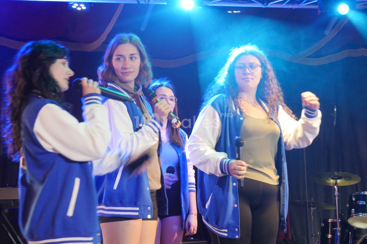 Quatre jeunes femmes en vestes bleues chantent sur scène, éclairées par des projecteurs, avec des micros en main.