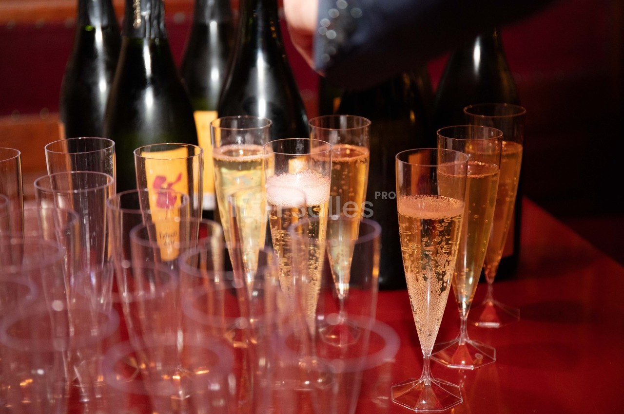 Flûtes de champagne remplies sur une table rouge, avec des bouteilles en arrière-plan.