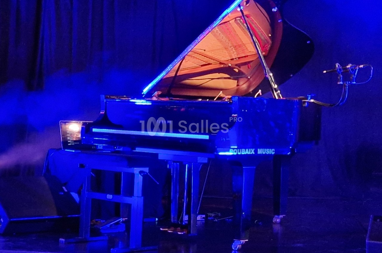 Piano à queue noir éclairé par des lumières bleues sur une scène sombre, avec un micro et un tabouret à proximité.