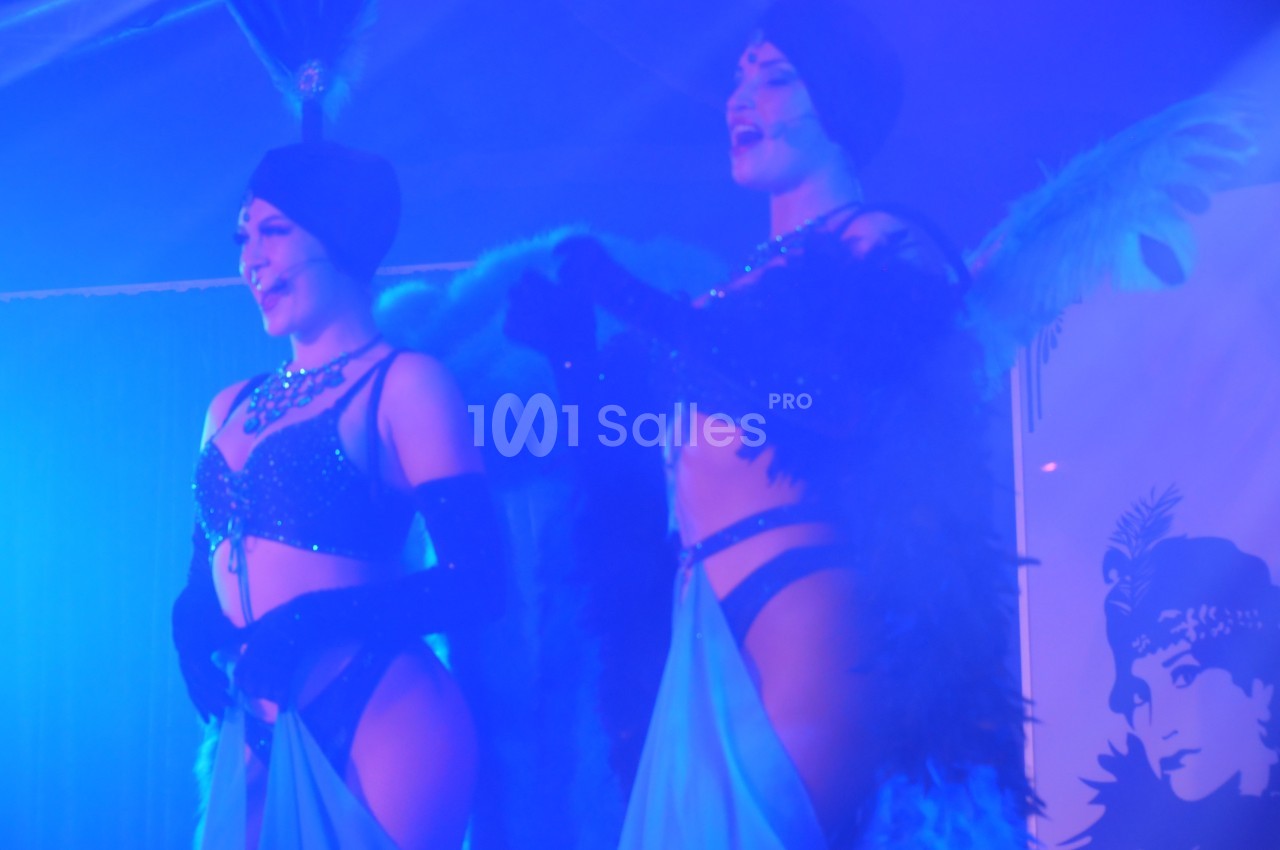 Deux artistes en costumes de cabaret avec plumes et paillettes se produisent sur une scène éclairée en bleu.