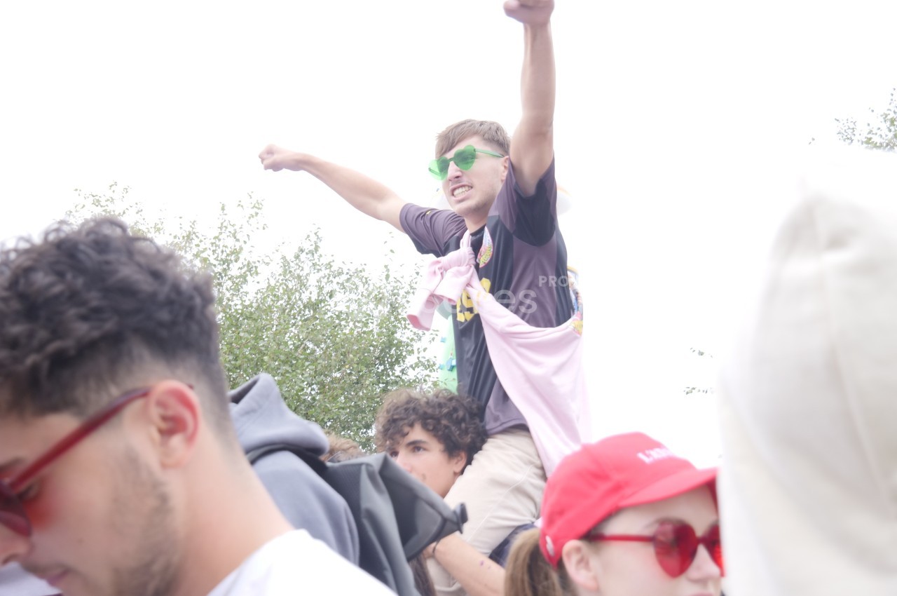 Un jeune homme portant des lunettes vertes lève les bras en l'air, assis sur les épaules d'une autre personne dans une foule.