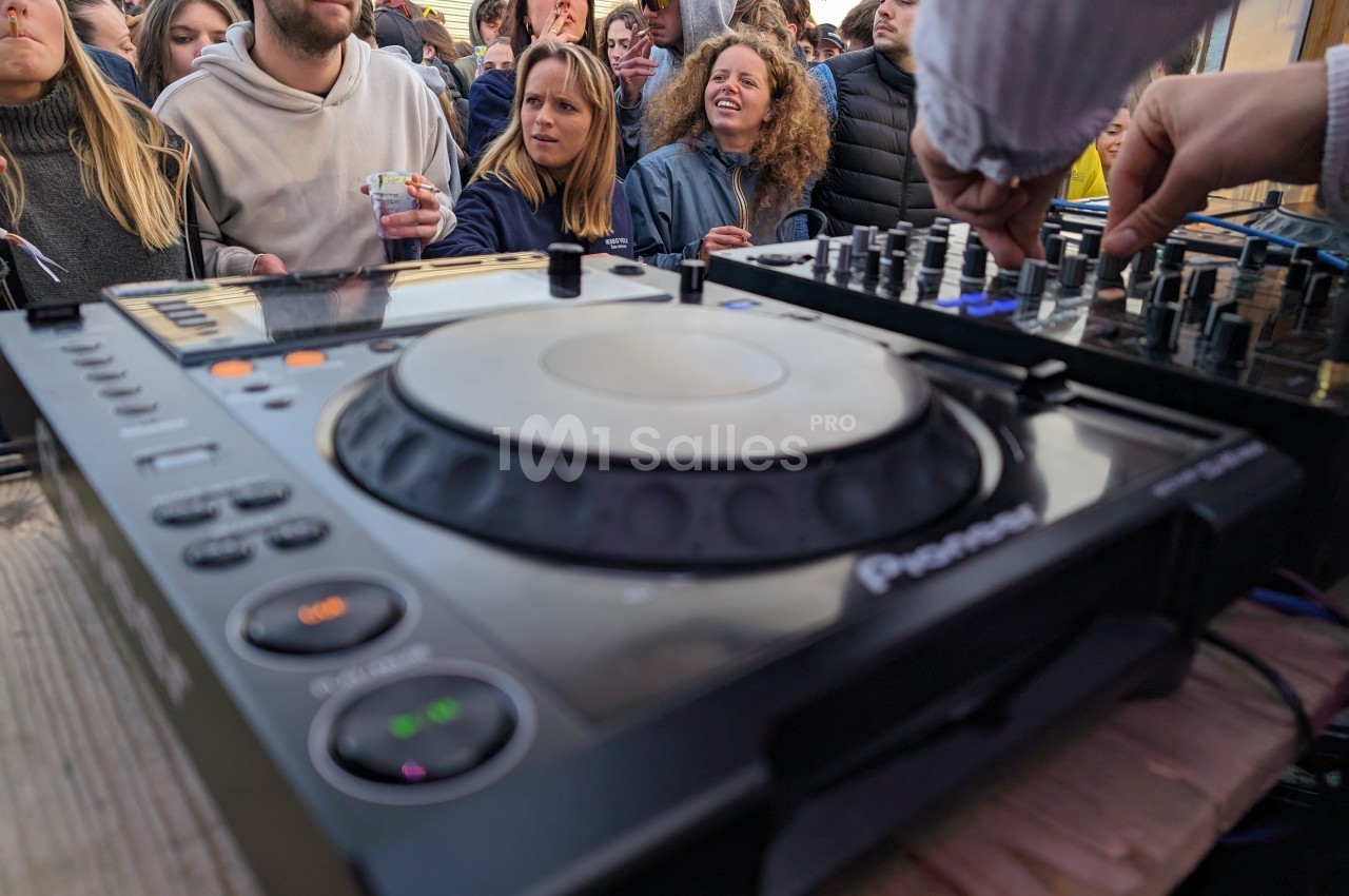 Vue rapprochée d'une table de mixage en usage lors d'un événement en plein air, avec un public en arrière-plan.