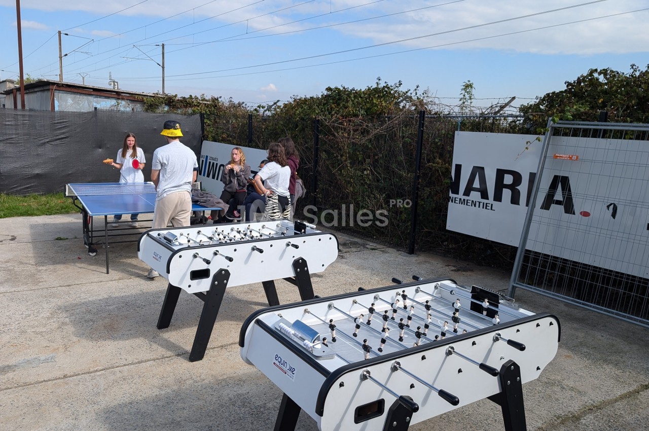 Deux babyfoots et une table de ping-pong en extérieur, avec des personnes discutant et jouant en arrière-plan.