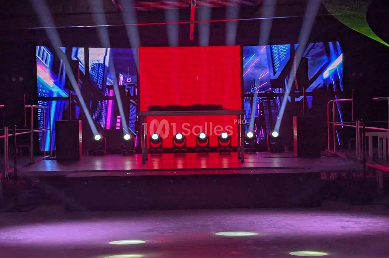 Scène de concert vide avec éclairage coloré, écran rouge central et équipements audio en arrière-plan.