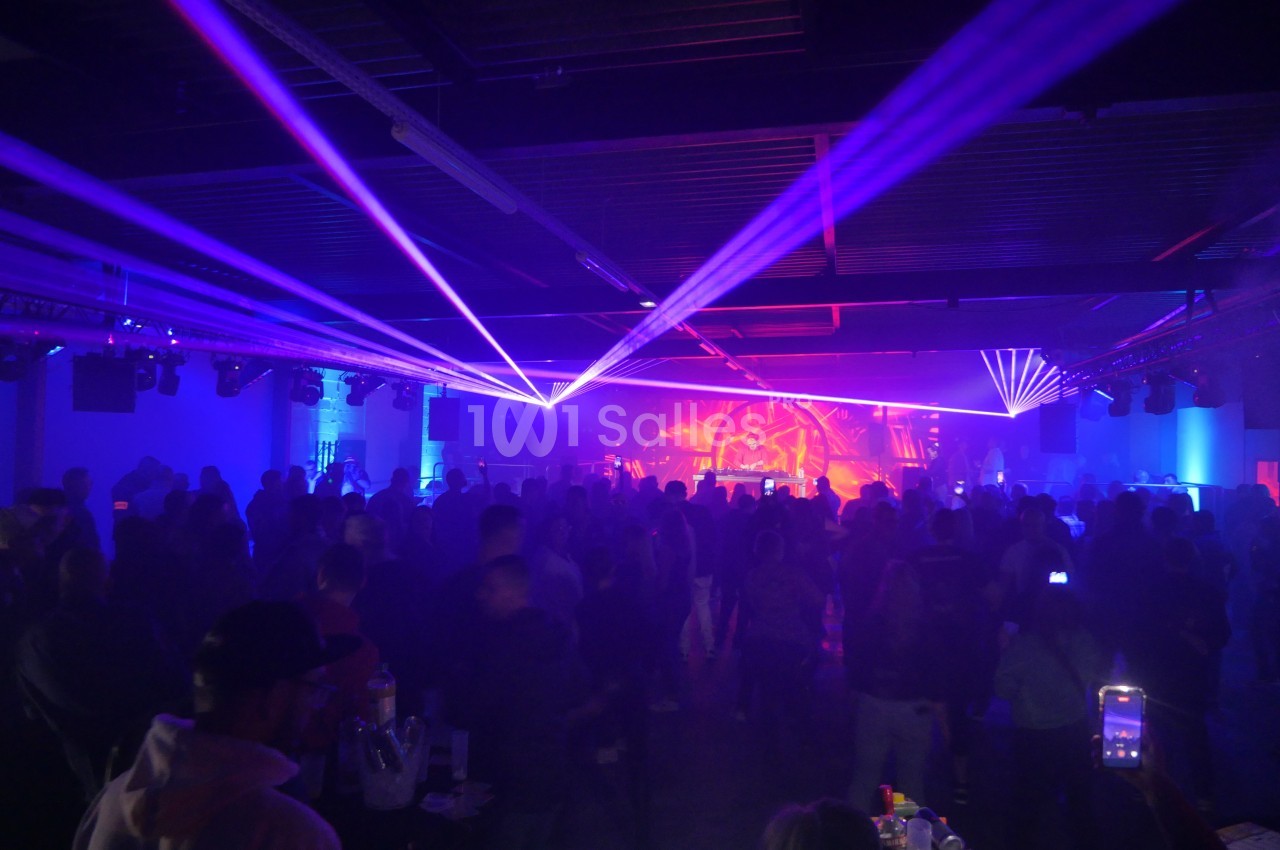 Salle sombre avec une foule debout, éclairée par des faisceaux lumineux colorés et une scène en arrière-plan.