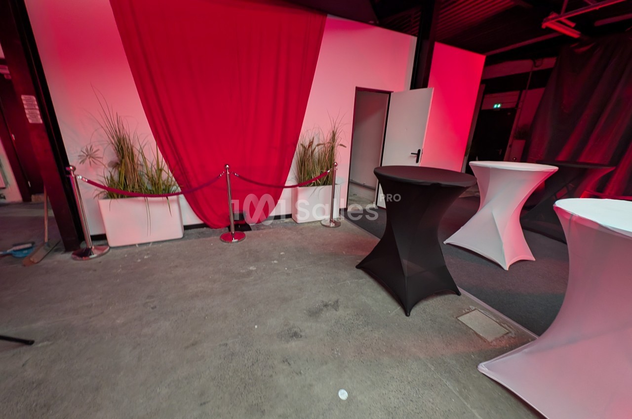 Salle avec nappes sur tables hautes, rideau rouge en fond, plantes en bacs blancs et sol en béton.