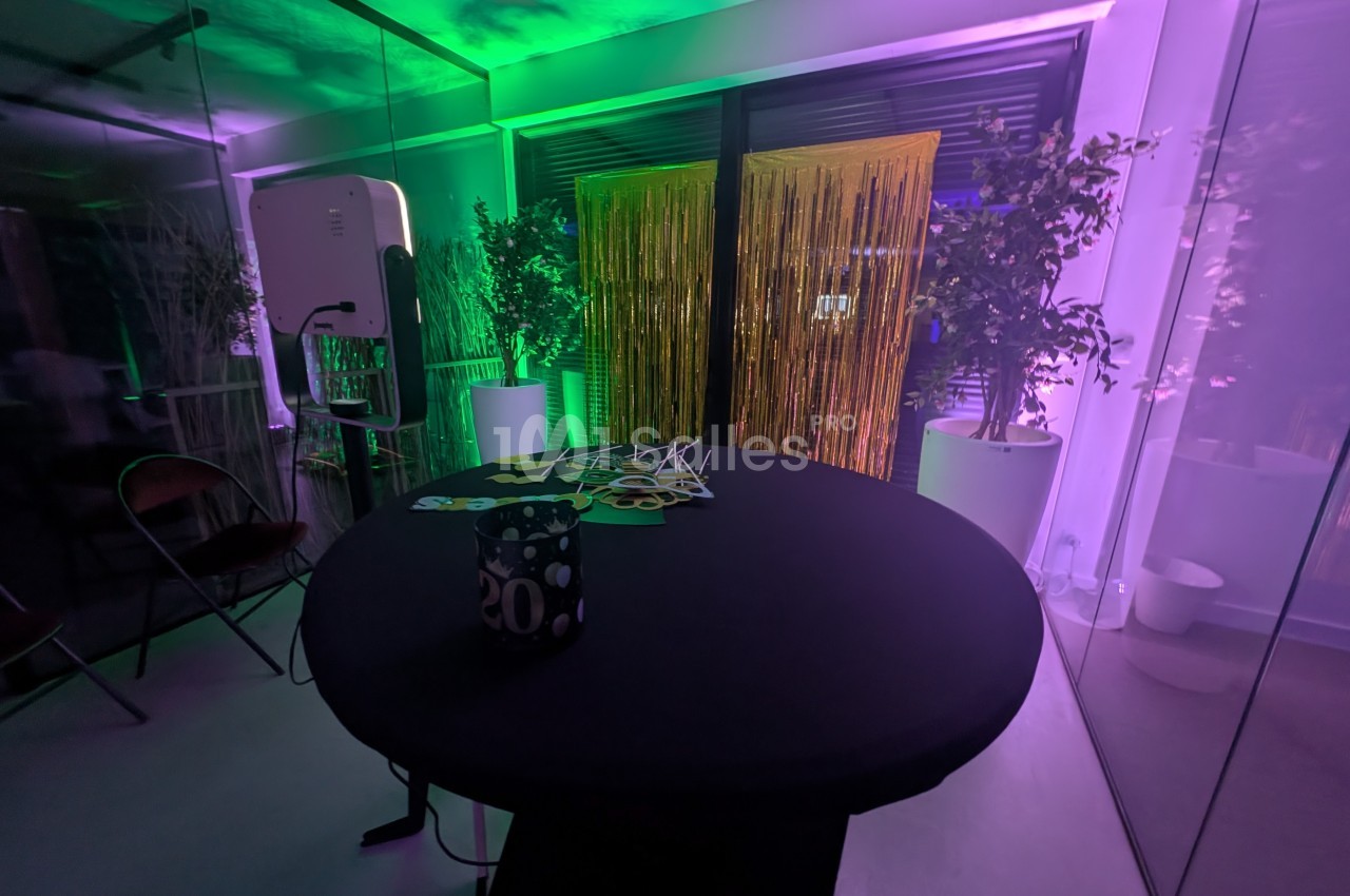 Salle décorée avec une table ronde noire, accessoires festifs, rideaux dorés et éclairage coloré vert et violet.
