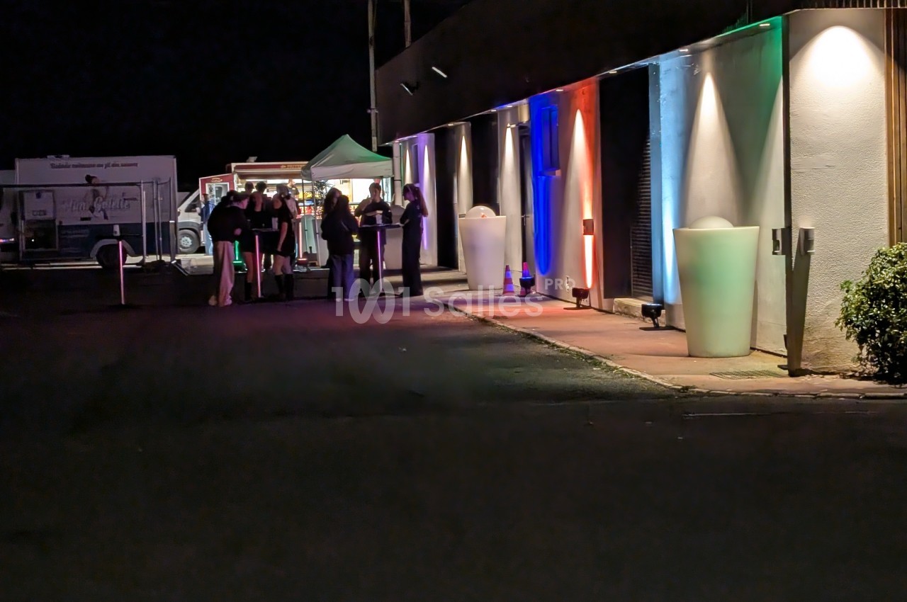 Groupe de personnes rassemblées près d'un stand éclairé, devant un bâtiment illuminé de lumières colorées, la nuit.