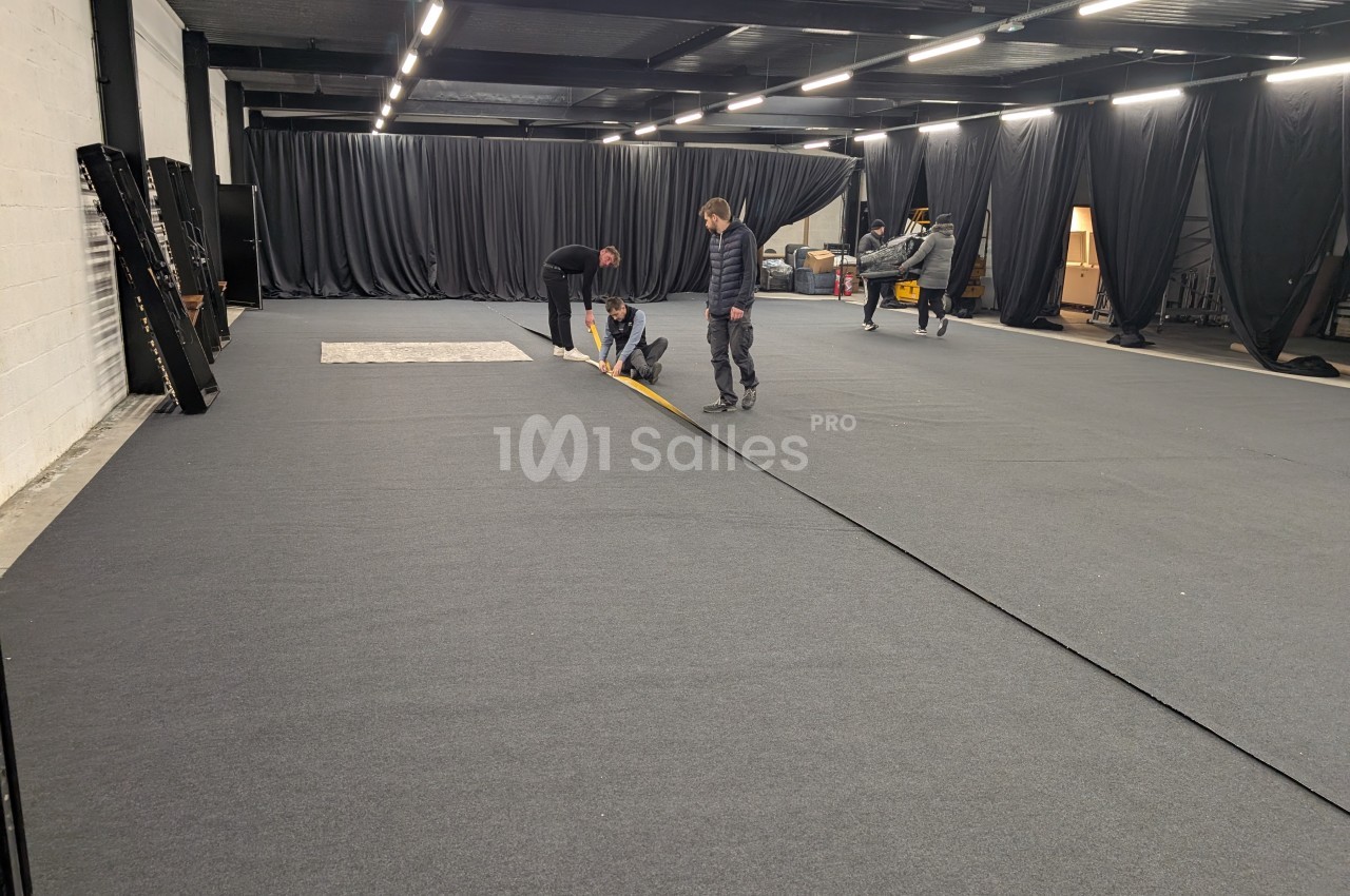 Des personnes installent un revêtement de sol gris dans un grand espace intérieur avec des rideaux noirs.