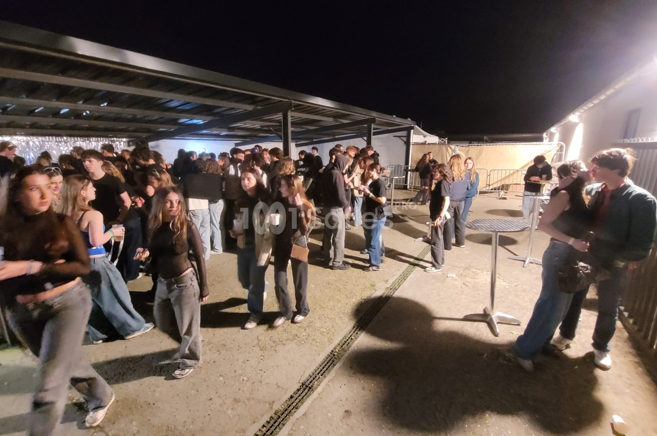 Groupe de personnes rassemblées en extérieur sous un abri, de nuit, certaines discutant ou tenant des boissons.
