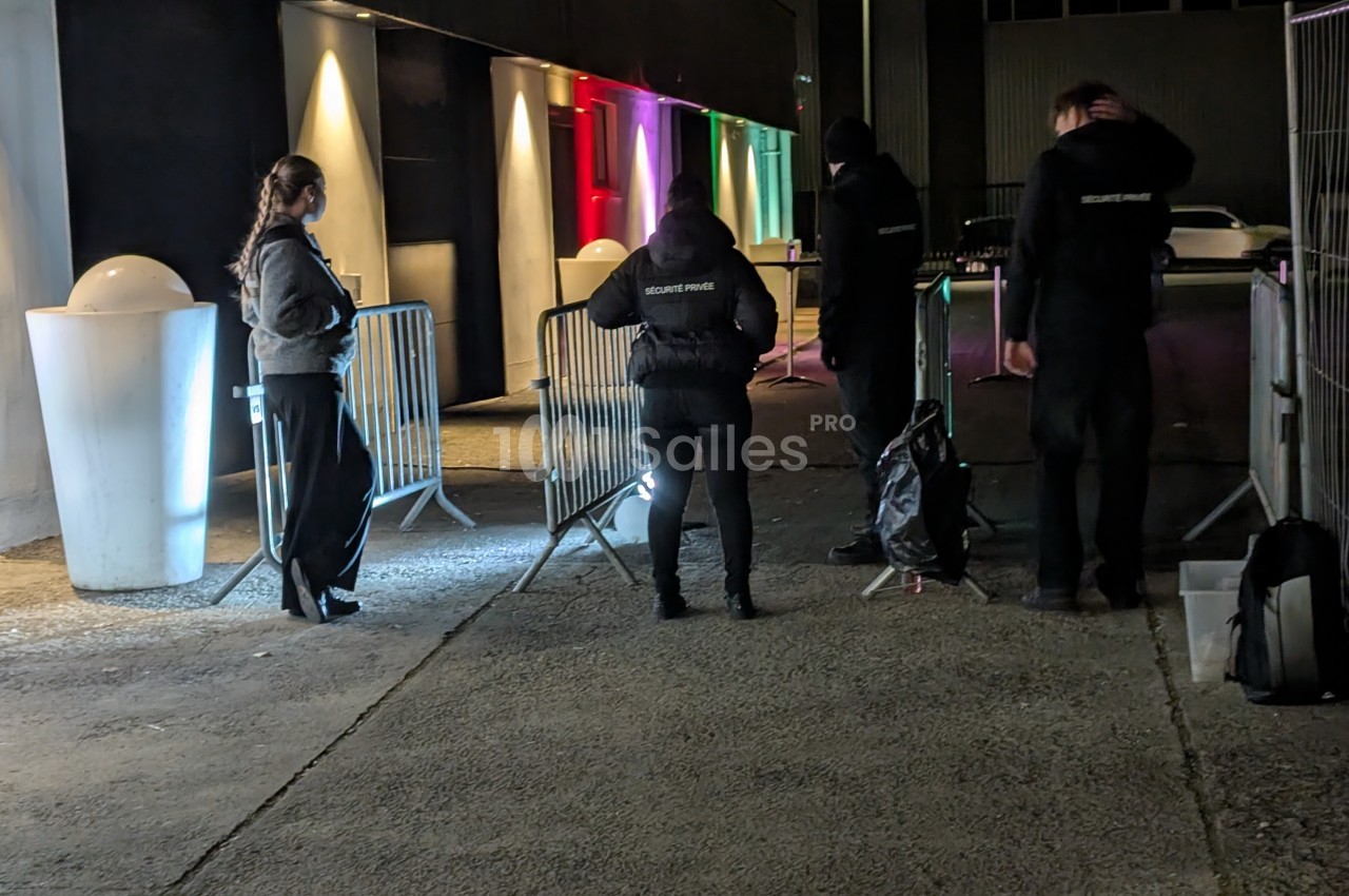 Des agents de sécurité surveillent une entrée extérieure de nuit, avec des barrières et un éclairage coloré en arrière-plan.