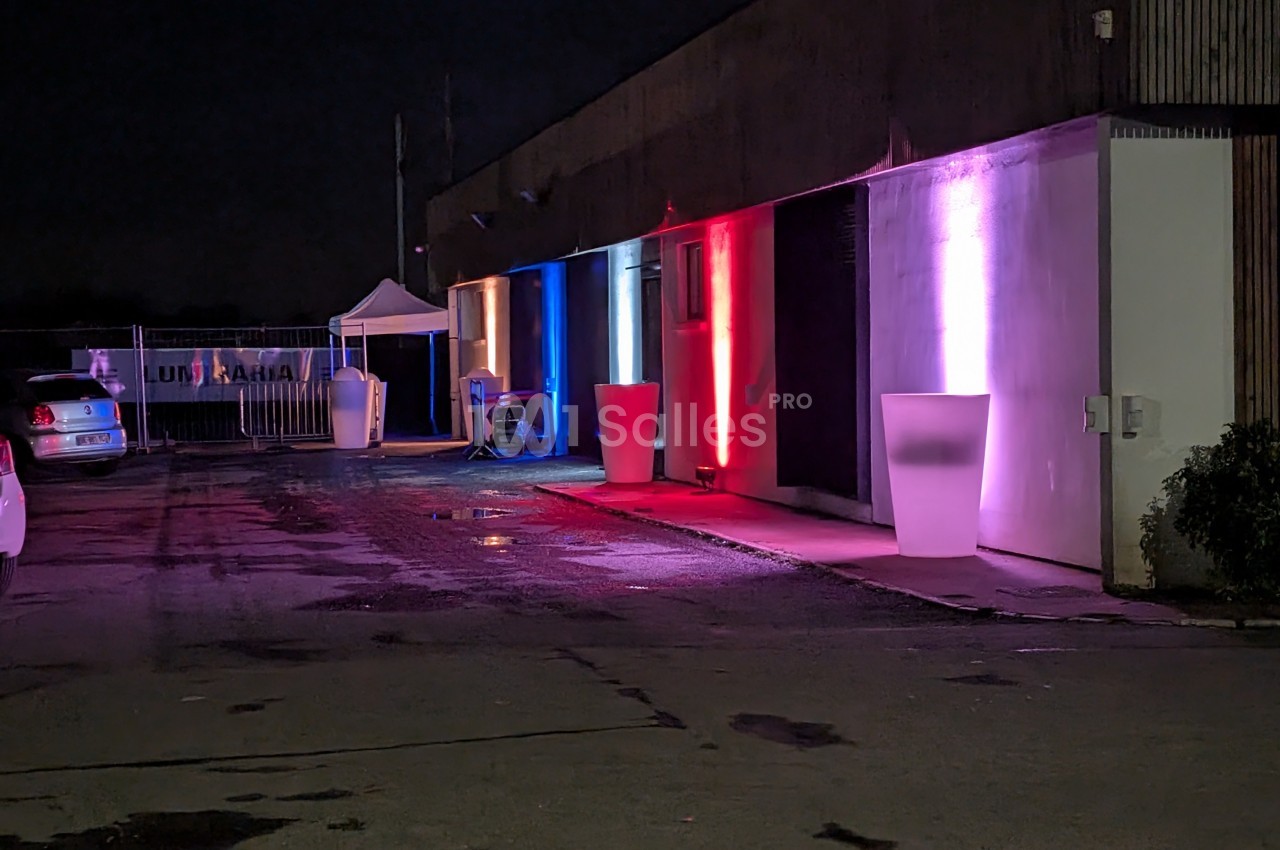 Façade d'un bâtiment éclairée par des lumières colorées rouge, bleue et blanche, avec des objets lumineux devant.