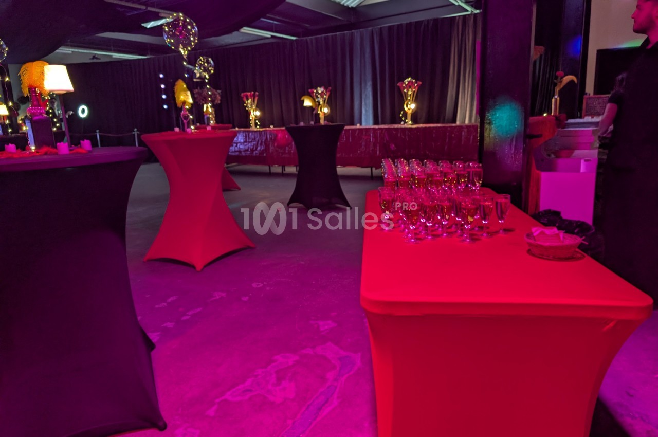 Salle de réception avec tables hautes drapées, éclairage coloré et verres disposés sur une table rouge.