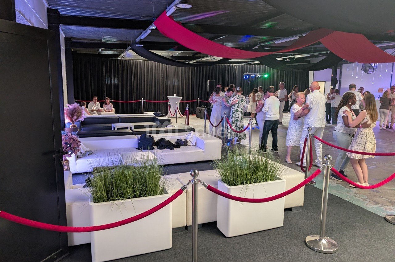 Salle de réception avec canapés blancs, plantes décoratives et invités debout ou discutant près des tables.
