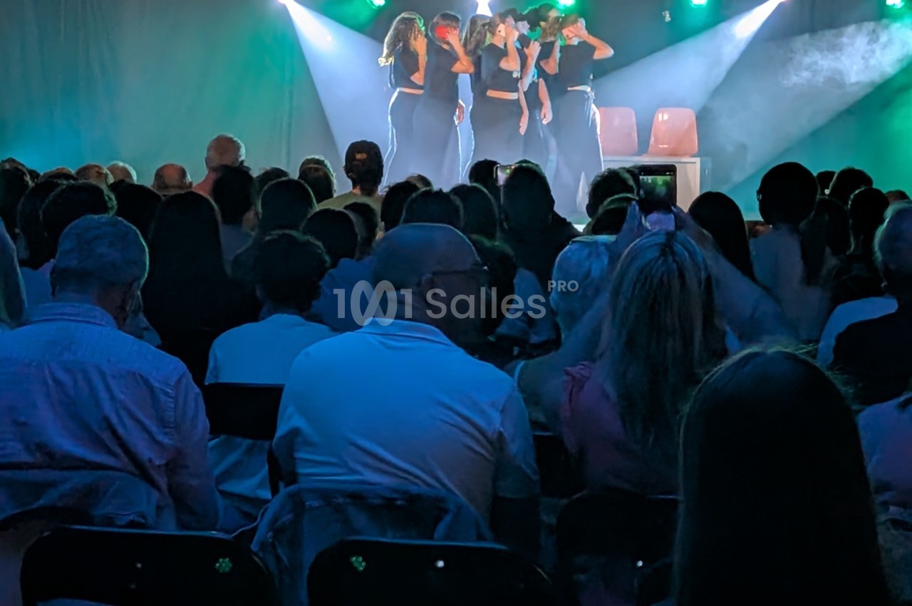 Un groupe de personnes sur scène sous des projecteurs, observé par un public assis dans une salle sombre.