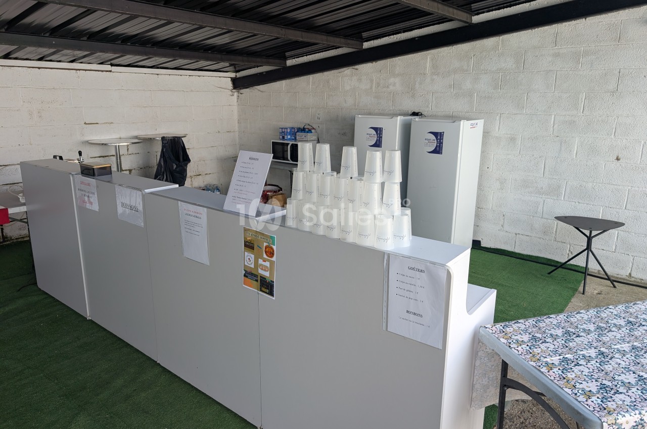 Comptoir blanc avec gobelets empilés, affiches et équipements de cuisine en arrière-plan sous un toit métallique.
