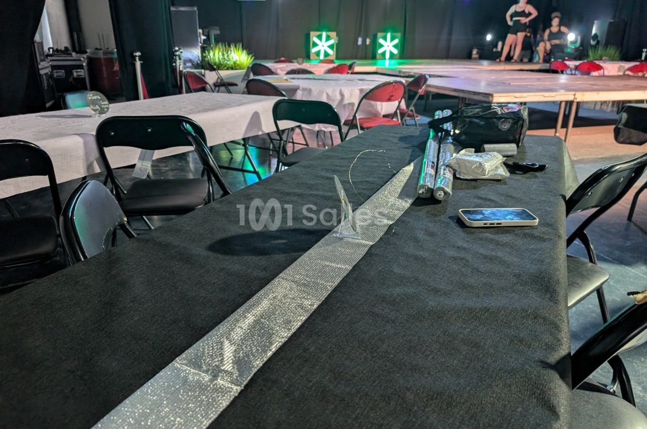 Table recouverte d'une nappe noire avec des décorations argentées, entourée de chaises dans une salle éclairée.