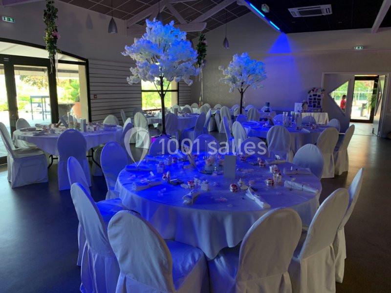 Salle de réception décorée avec des tables rondes, nappes blanches, chaises houssées et arbres artificiels illuminés.