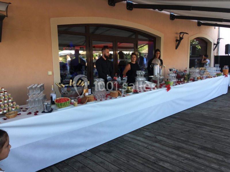 Table de buffet garnie de boissons, verrines, fruits et coupes, avec trois serveurs en arrière-plan.