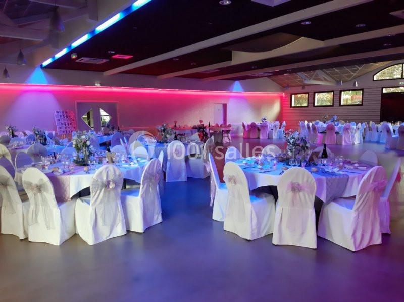 Salle de réception décorée avec des tables rondes dressées, nappes blanches et chaises couvertes, éclairage coloré.