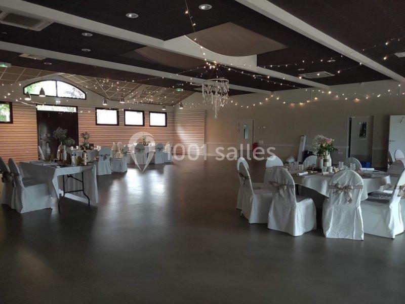 Salle de réception décorée avec des tables rondes, des chaises couvertes et des guirlandes lumineuses suspendues.