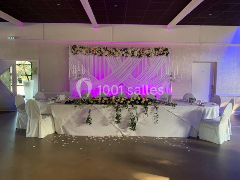 Table décorée pour un événement, ornée de fleurs blanches et roses, avec un fond lumineux et des chaises habillées.