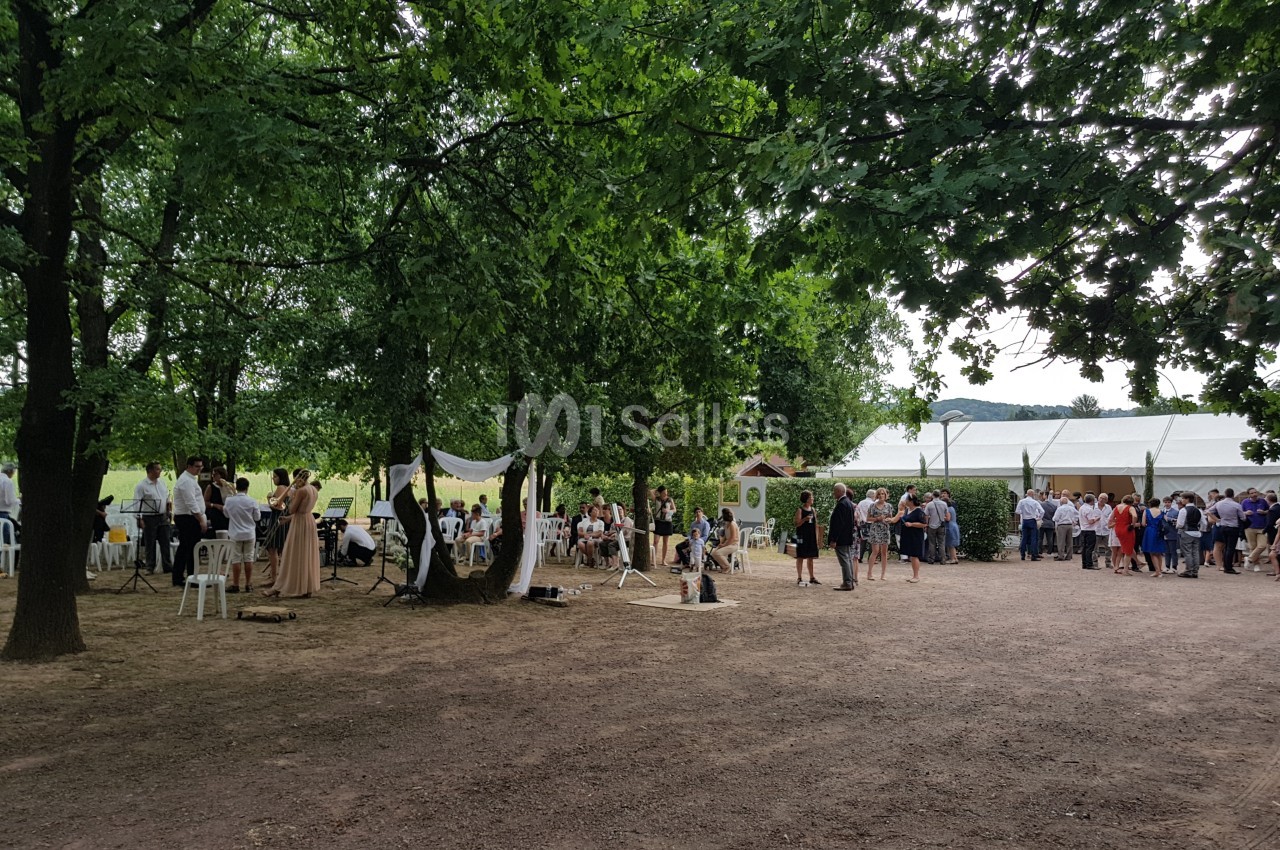 Groupe de personnes rassemblées en plein air sous des arbres, avec des chaises et une arche décorative en arrière-plan.