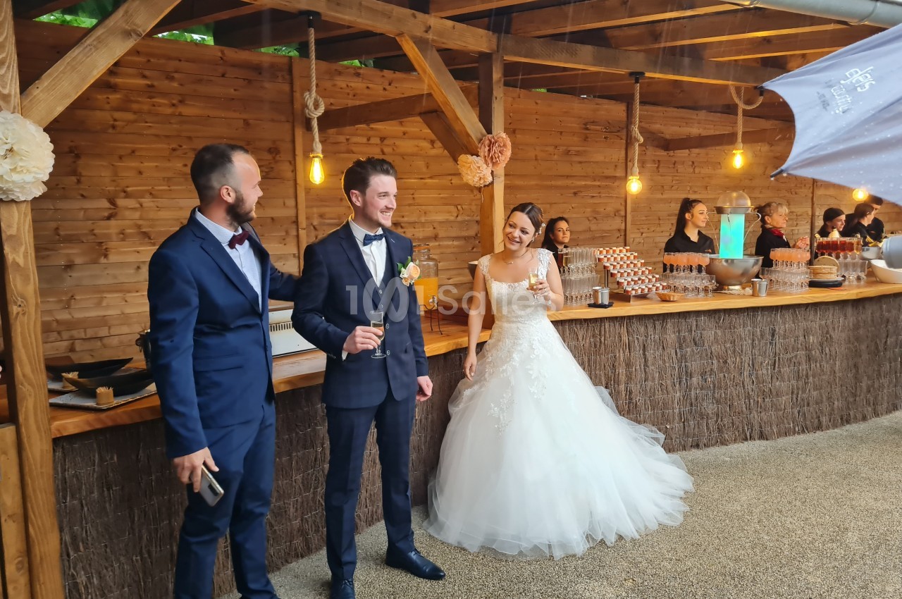 Un couple de mariés et un invité discutent devant un bar en bois décoré lors d'une réception en extérieur.