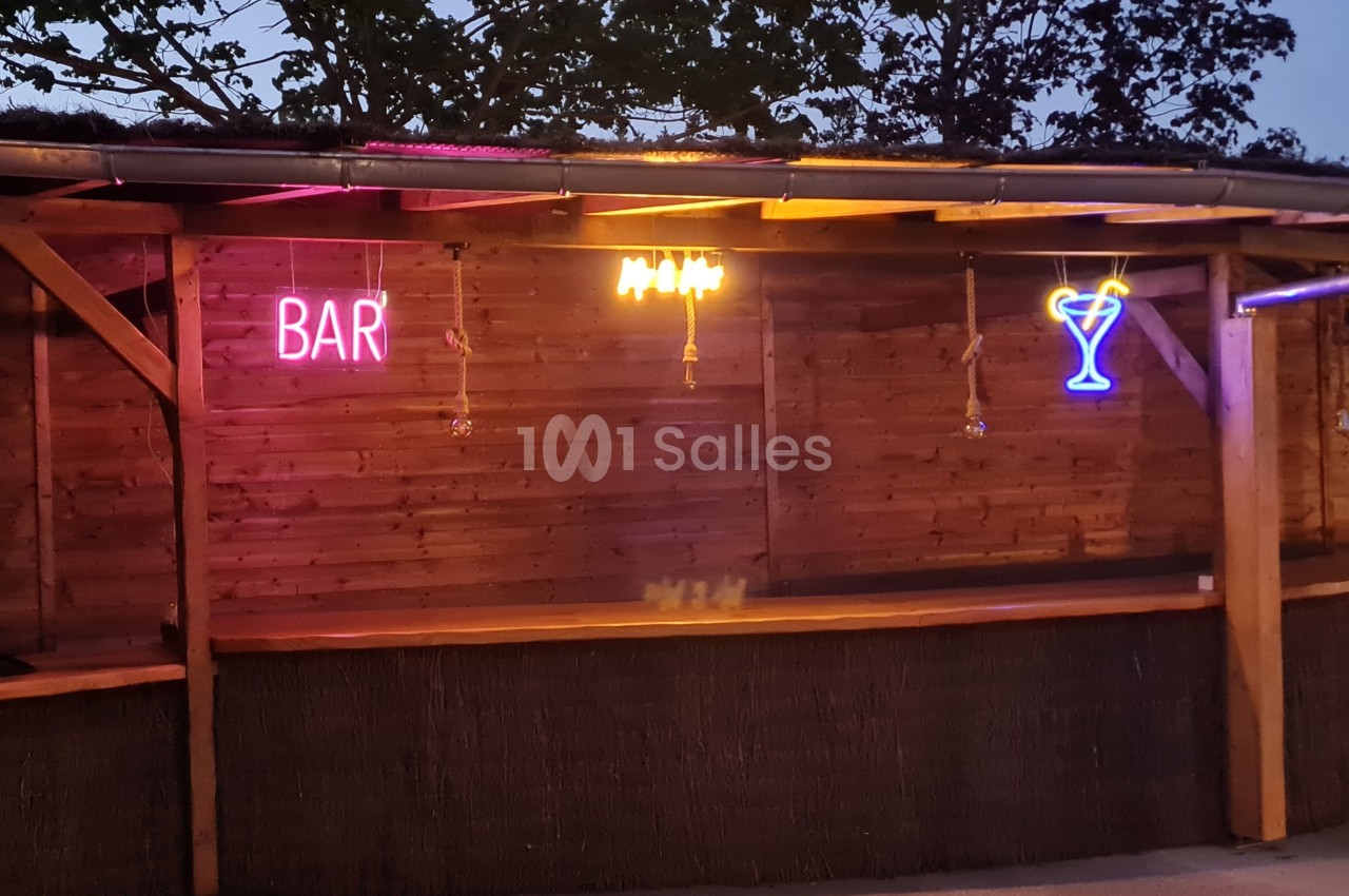 Bar extérieur en bois avec des néons colorés représentant le mot ’BAR’ et un cocktail, éclairé en soirée.