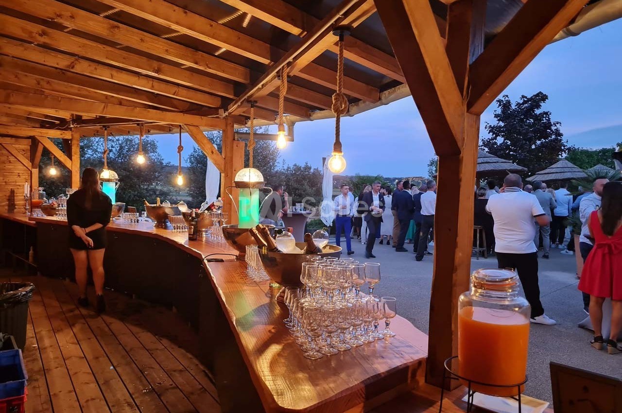 Bar en bois éclairé avec des boissons et des verres, donnant sur une terrasse où des personnes sont rassemblées en soirée.