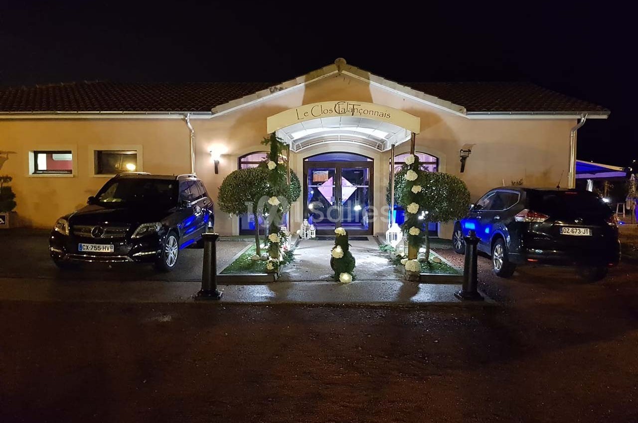 Façade éclairée d'un bâtiment avec deux voitures garées devant, entourée de décorations végétales.