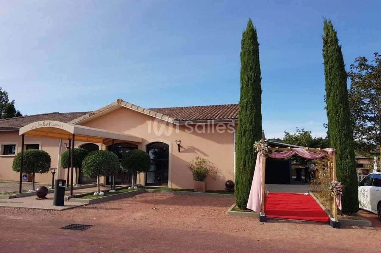 Façade d'un bâtiment avec entrée décorée, tapis rouge et arche fleurie, entouré de cyprès et d'arbustes taillés.