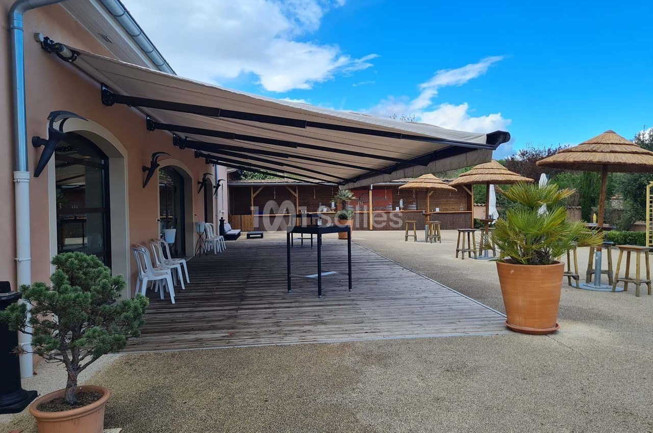 Terrasse avec auvent déployé, tables hautes, chaises blanches et plantes en pots, sous un ciel partiellement dégagé.