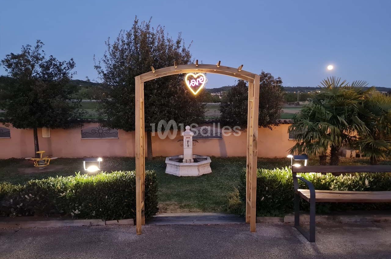 Arche en bois avec un néon en forme de cœur, devant une fontaine dans un jardin éclairé au crépuscule.
