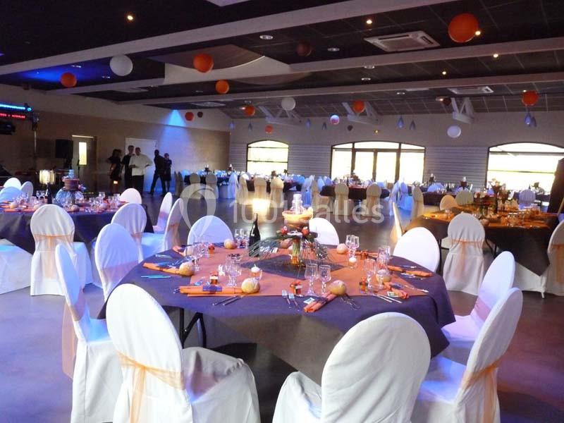 Salle de réception décorée avec des tables rondes dressées, nappes violettes et chaises couvertes de housses blanches.