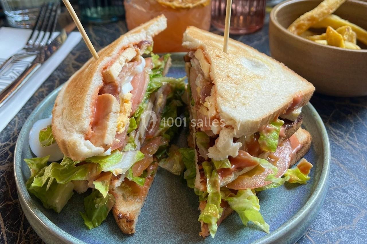 Sandwich club garni de laitue, tomate, poulet et bacon, présenté sur une assiette avec des frites en arrière-plan.