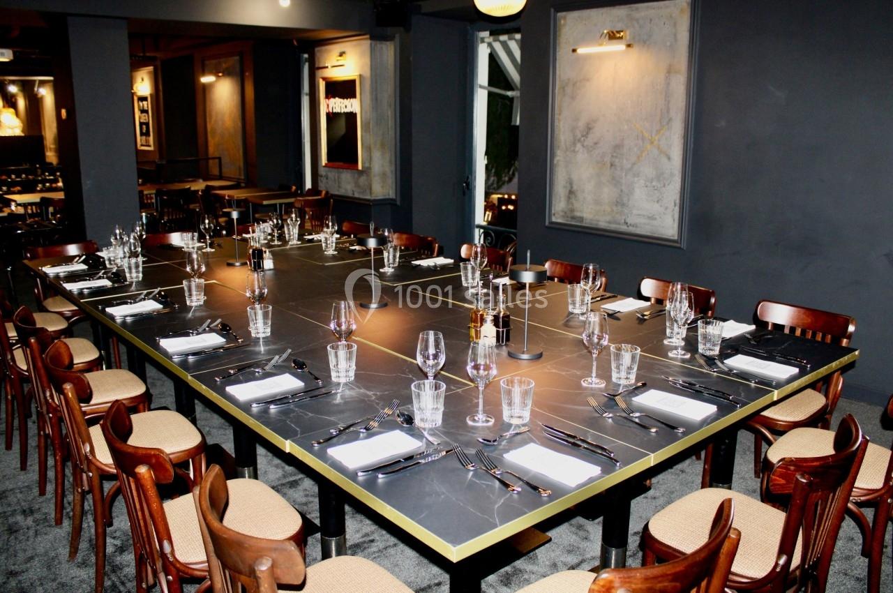 Grande table rectangulaire dressée avec couverts, verres et serviettes, entourée de chaises en bois dans une salle sombre.