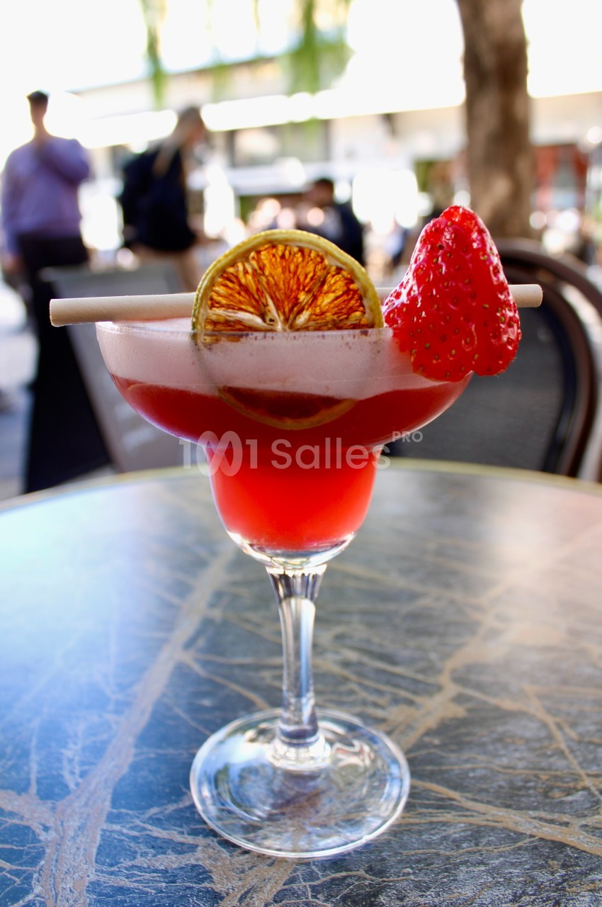 Verre de cocktail rouge garni d'une tranche d'orange séchée et d'une fraise, posé sur une table en extérieur.