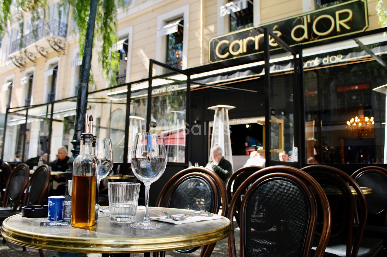 Terrasse d'un café avec des tables et chaises en bois, verres et bouteille d'huile sur une table, devant une façade vitrée.