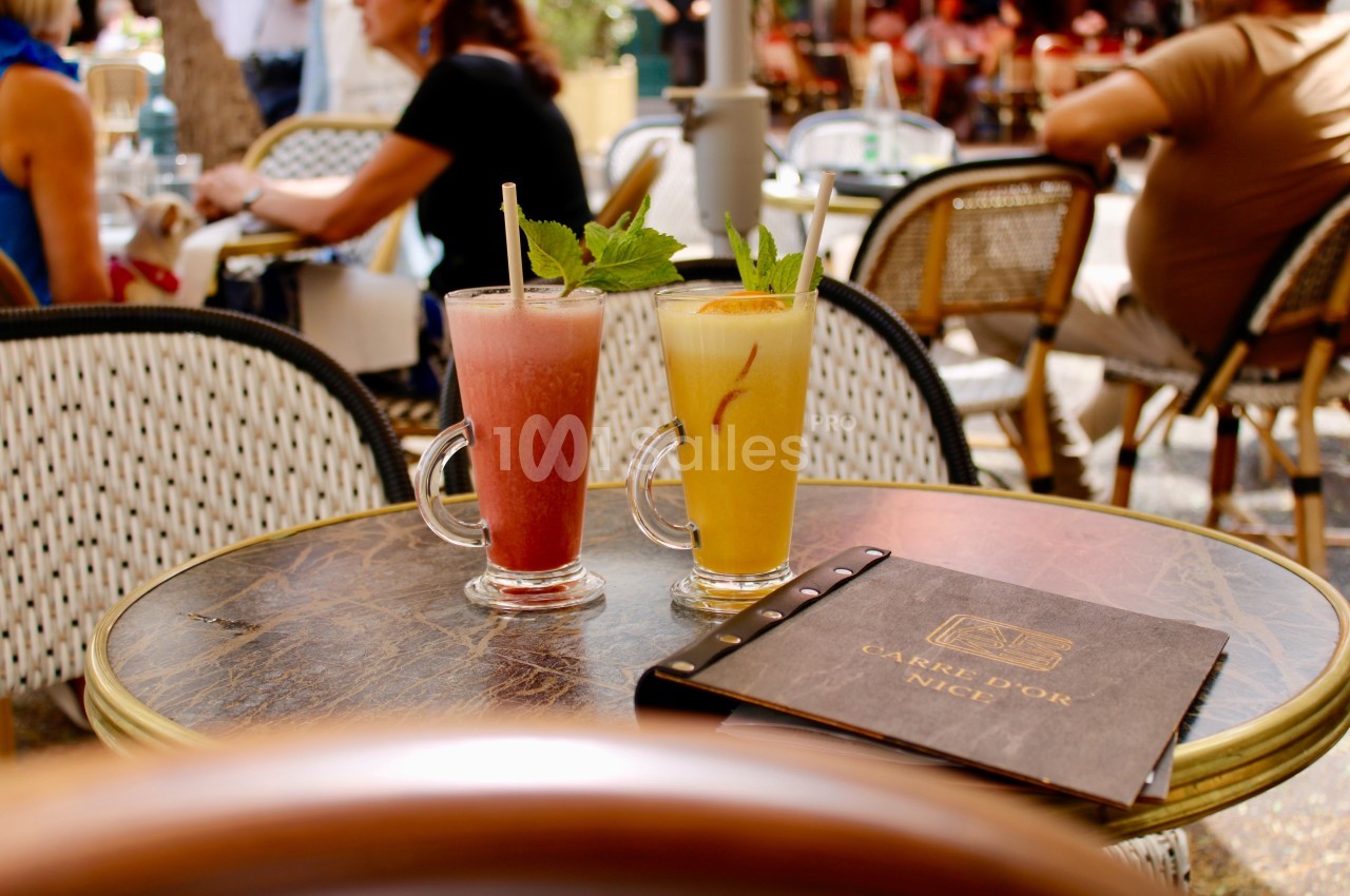 Deux verres de cocktails colorés sur une table en terrasse, entourée de chaises en osier et d'une ambiance animée.