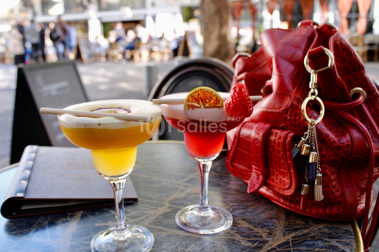 Deux cocktails colorés sur une table en terrasse, accompagnés d'un sac rouge et d'un menu en arrière-plan.