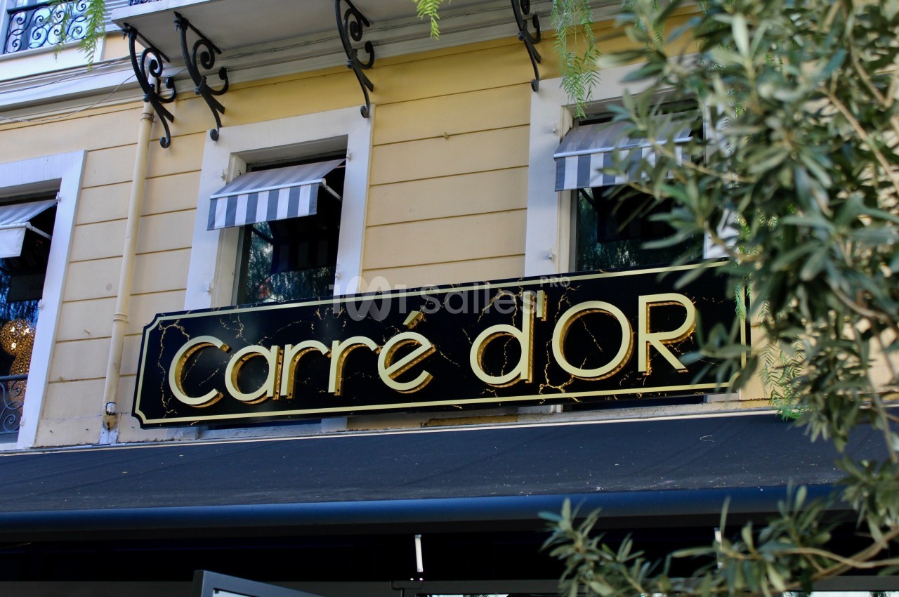Enseigne noire et dorée avec l'inscription ’Carré d'Or’ sur la façade d'un bâtiment jaune avec des stores rayés.