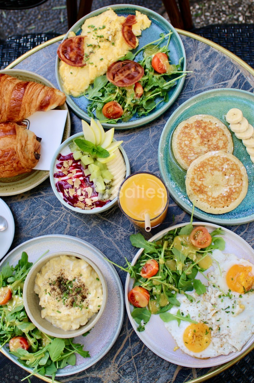 Petit-déjeuner varié avec croissants, œufs, pancakes, fruits, salade, café et jus d'orange sur une table ronde.