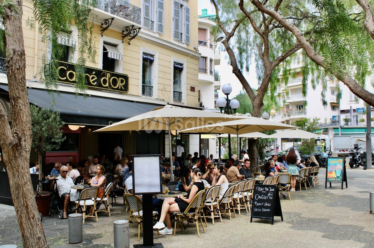 Terrasse animée d'un café en plein air avec des clients assis sous des parasols, entourée d'arbres et de bâtiments.