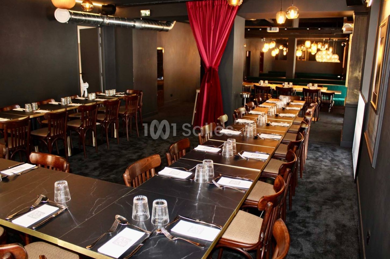 Salle de restaurant élégante avec longues tables noires, chaises en bois, rideaux rouges et éclairage chaleureux.