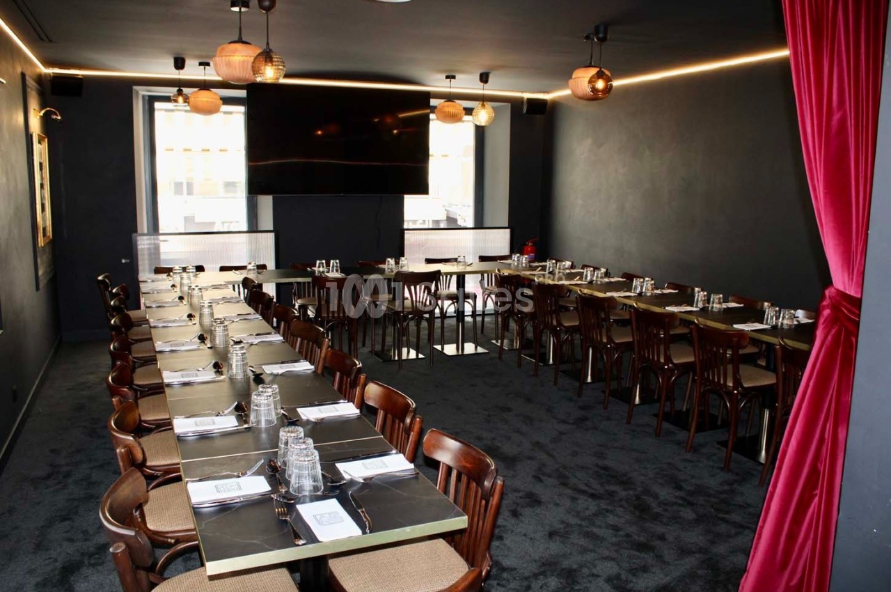 Salle de restaurant sombre avec deux longues tables dressées, chaises en bois et rideaux rouges encadrant l'entrée.