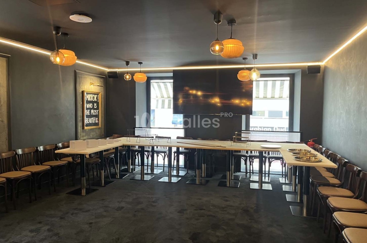 Salle de réunion moderne avec tables disposées en U, chaises en bois, éclairage suspendu et écran mural.